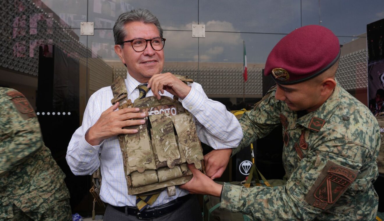 Ricardo Monreal ajusta un chaleco táctico con ayuda de un militar durante una exhibición.